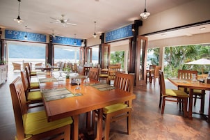 A dining area with wooden tables and chairs, large windows with ocean views, and ceiling fans.