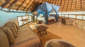 Ein Zimmer mit einem Strohdach, einem Himmelbett, einem Sofa, einem kleinen Tisch und einer Steinwand.