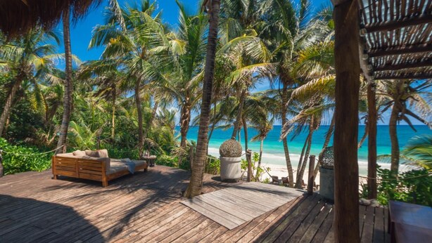 Eine Holzterrasse mit einer Couch und einem Tisch, umgeben von Palmen und mit Blick auf einen Strand.