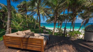 Eine Holzterrasse mit einem Bett und einem kleinen Tisch, umgeben von Palmen und mit Blick auf einen Strand.