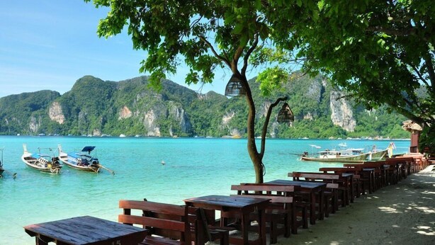 A beachside area with wooden benches and tables, boats on the water, and a mountainous backdrop. - Phi Phi Rimlay Resort (Phi Phi Islands)