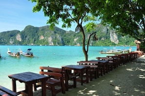 A beachside area with wooden benches and tables, boats on the water, and a mountainous backdrop. - Phi Phi Rimlay Resort (Phi Phi Islands)