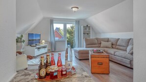 A cozy loft apartment with a high ceiling, a large window with curtains, a beige sofa, a wooden chest, and a dining table with bottles.