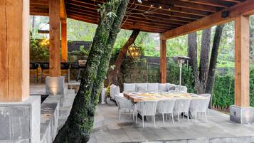 An outdoor patio with a wooden pergola, stone flooring, and a round table with white chairs.