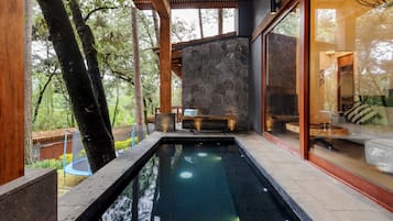 A modern outdoor pool area with a stone wall, wooden pergola, and a glass enclosure with a sofa and table.