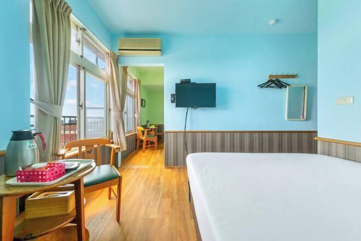 Een hotelkamer met een bed, een bureau met een waterkoker, een stoel, een televisie en een raam met gordijnen.