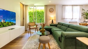 A modern living room with a green sofa, a flat-screen TV, a small round table, a chair, and a large window with a view of greenery.