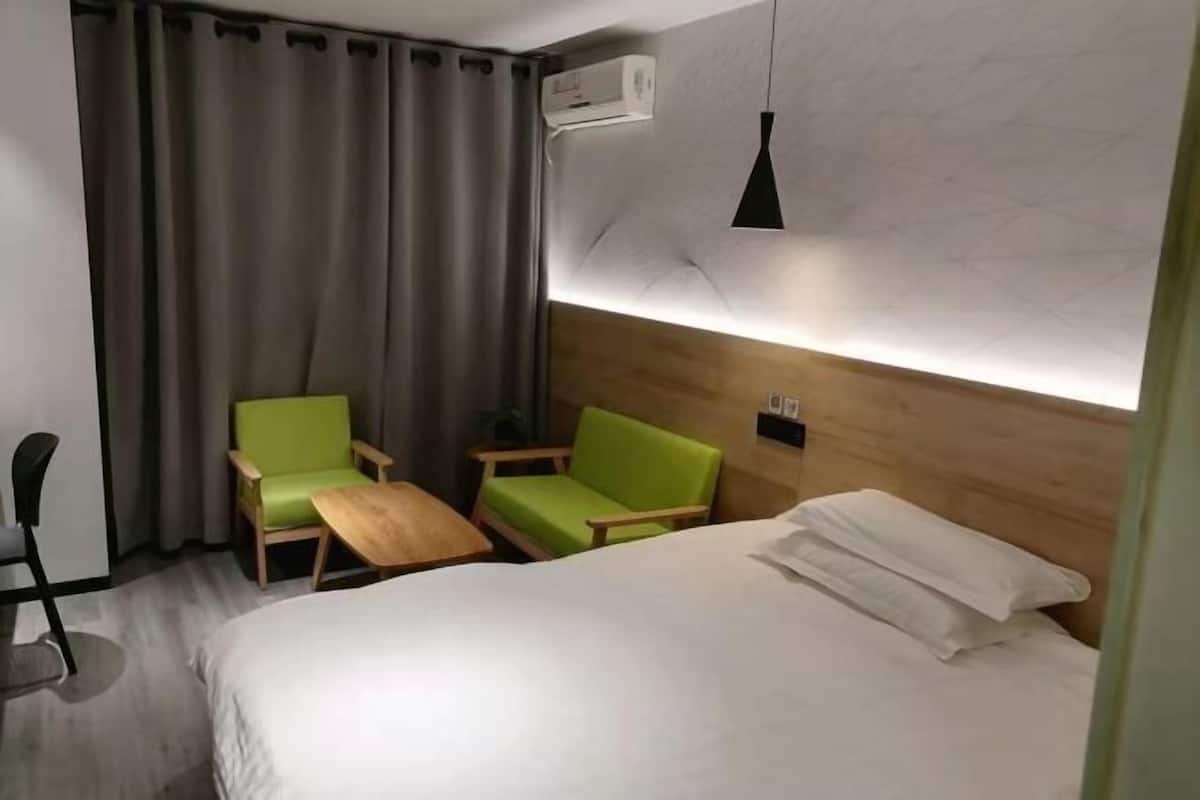 A hotel room with a bed, two green chairs, a small wooden table, a black pendant light, and a wall-mounted air conditioner.