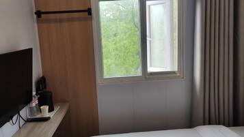 Habitación de hotel con una cama, ventana con cortinas, pared de madera y repisa con cafetera y taza.