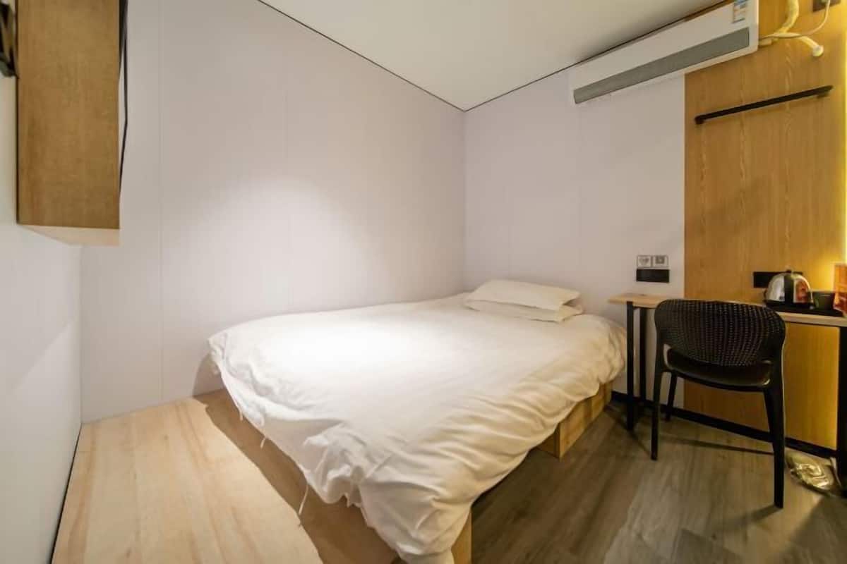 A small, single-bed room with a wooden floor, a desk with a chair, and a wall-mounted air conditioner.