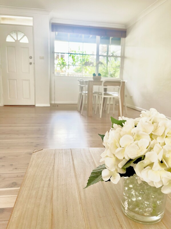 A room with a wooden floor, a white door, a dining table with chairs, and a window with a view of greenery.