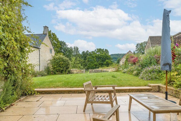 A patio with a wooden table and chairs, a stone wall, and a garden with a lawn and trees. - The Mill House Beautiful 4BD Family Home (Stroud)