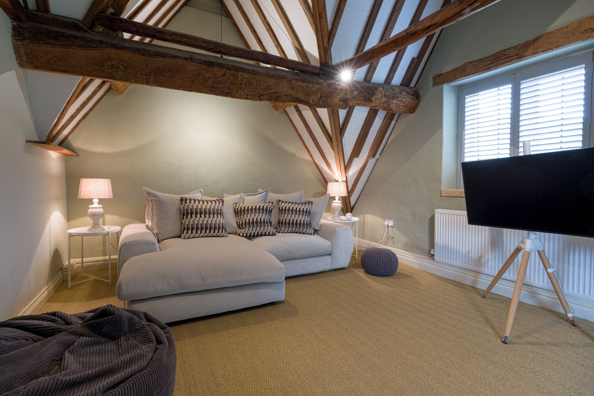 A cozy bedroom with a large bed, a TV on a tripod, a lamp, and a window with blinds.