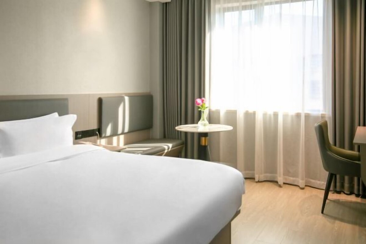 A hotel room with a large bed, a small table with a vase of flowers, a chair, and a view of the city through sheer curtains.