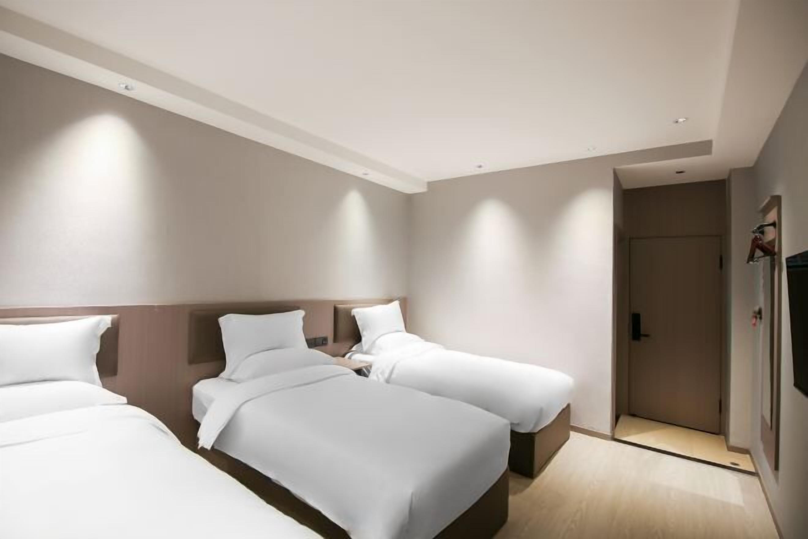 Habitación de hotel con tres camas dispuestas en fila, cabecera de madera y puerta que da a un pasillo.