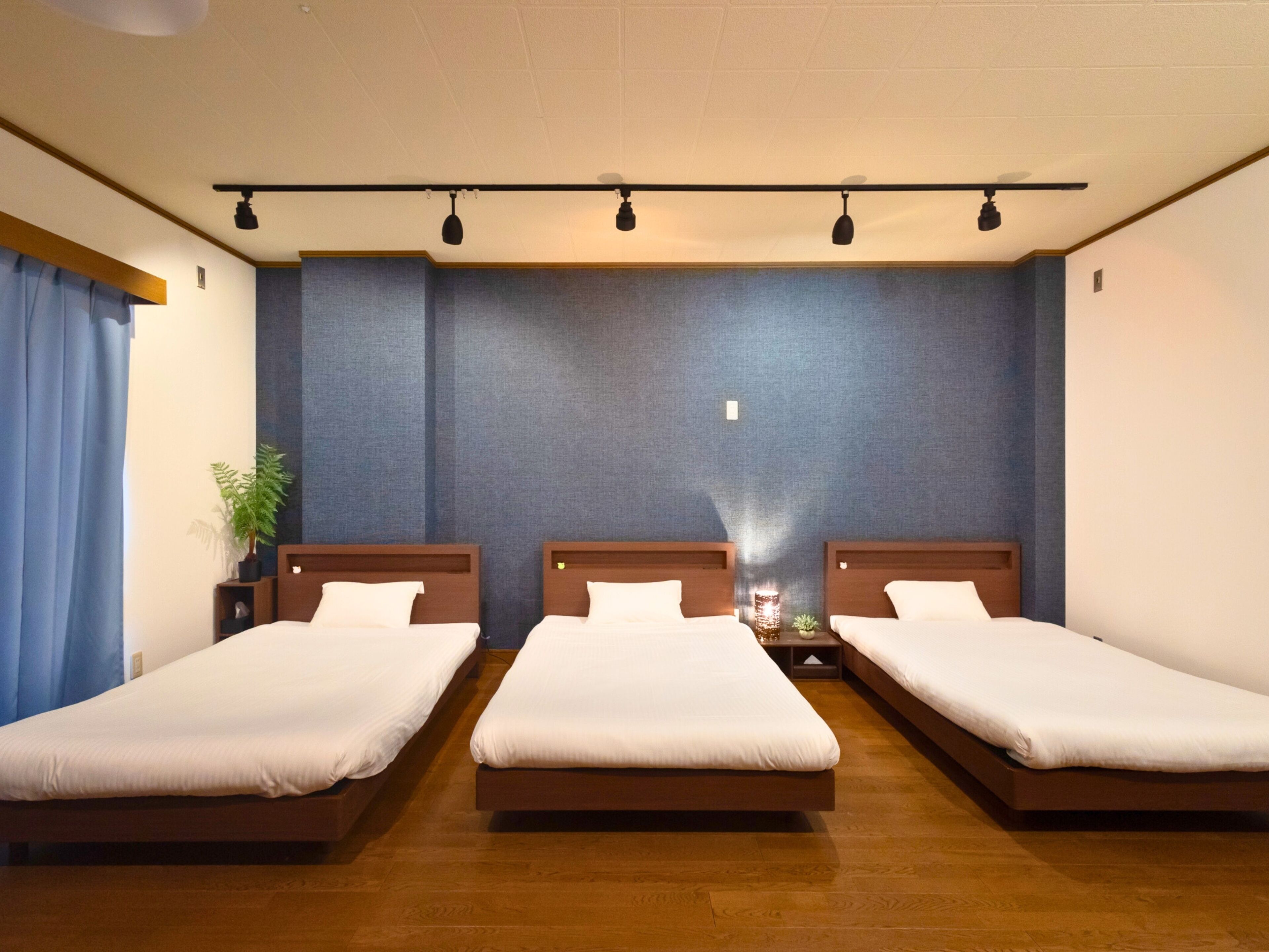 Une chambre d’hôtel avec trois lits simples, un plancher en bois, un mur bleu à texture et un plafond doté d’un éclairage encastré.