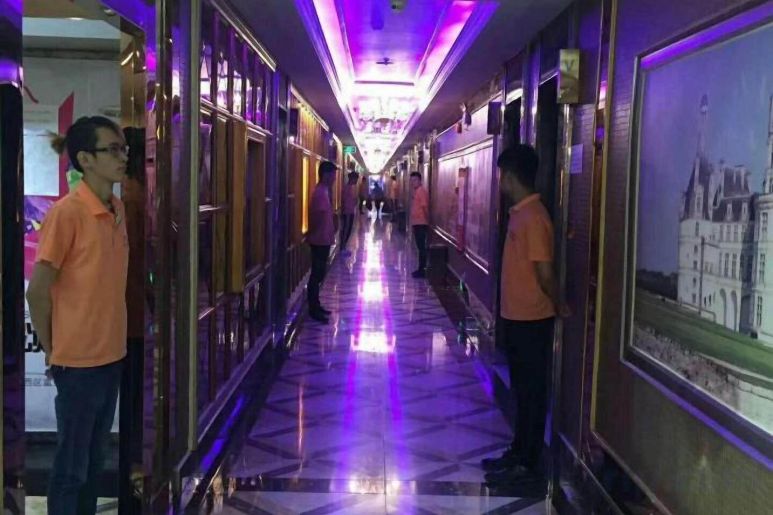 A long corridor with a reflective floor, framed pictures on the walls, and a person standing at the end.