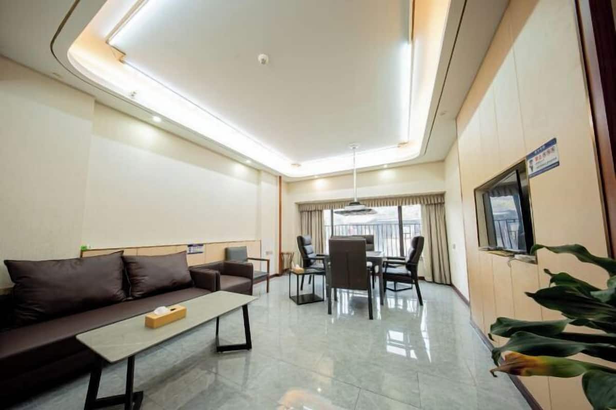 Een moderne hotelkamer met een bank, een klein tafeltje, een zithoek met stoelen, een aan de muur bevestigde televisie en een groot raam met gordijnen.