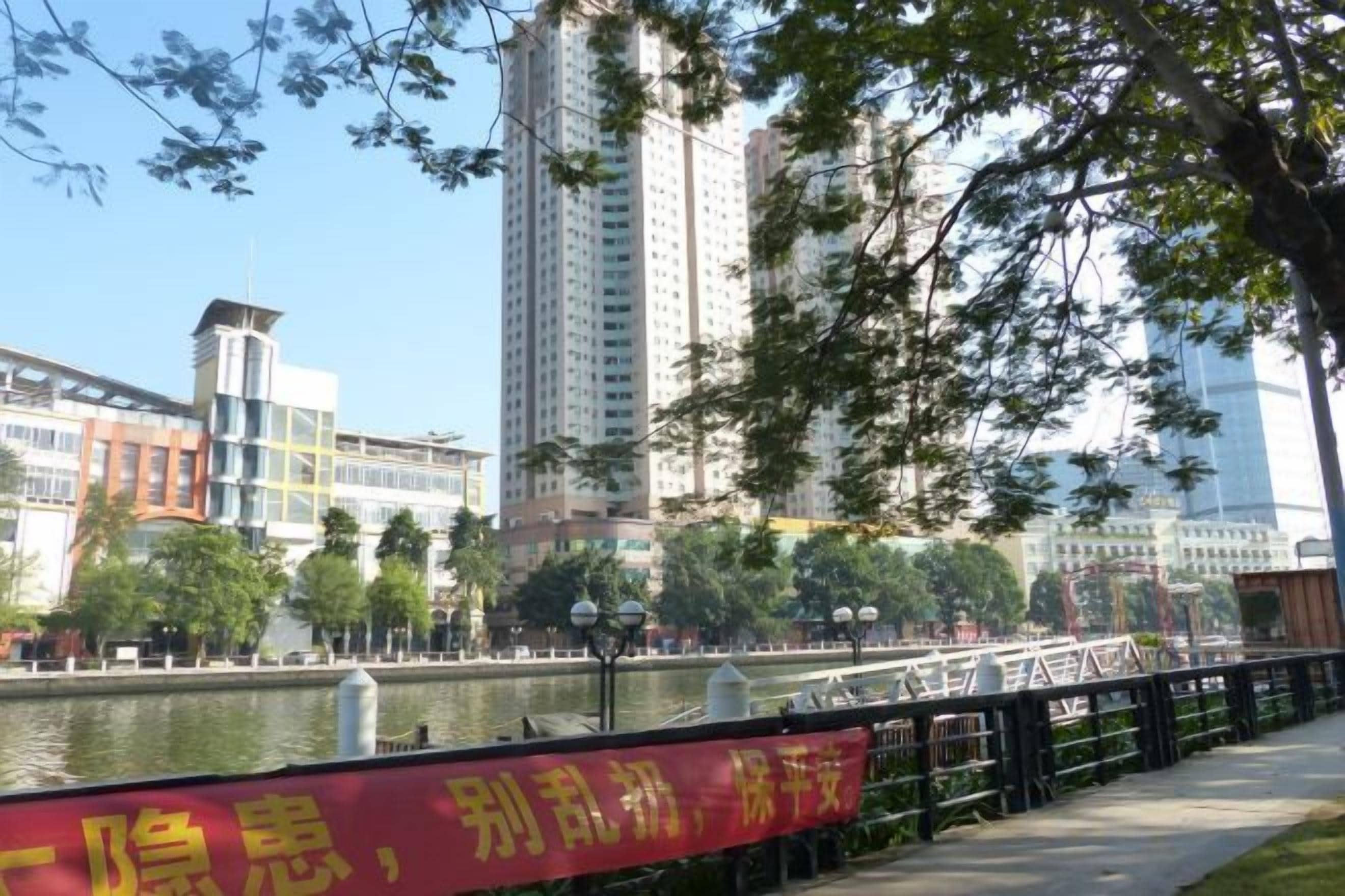 A riverside walkway with a red banner, a bridge, and high-rise buildings in the background.