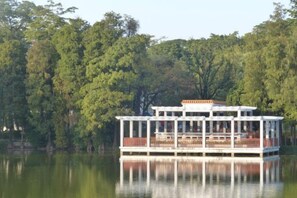 A stationary houseboat with a covered deck and multiple windows, surrounded by calm water and dense trees.