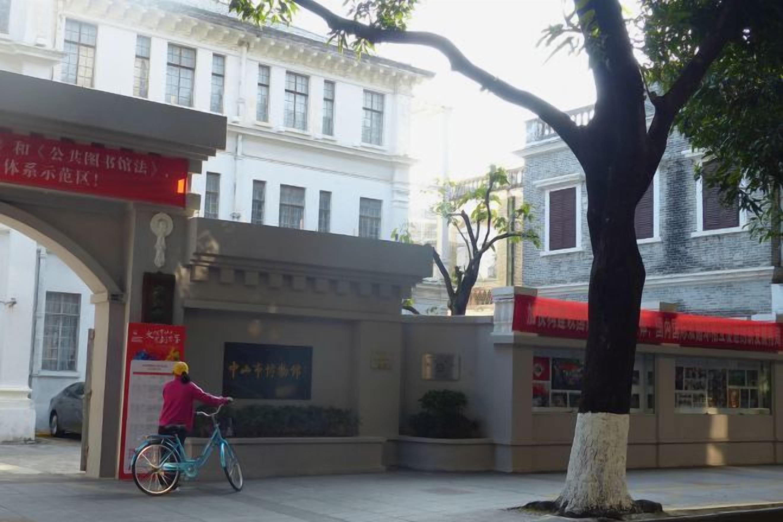 A person riding a bicycle past a building with a red sign and a tree in the foreground.