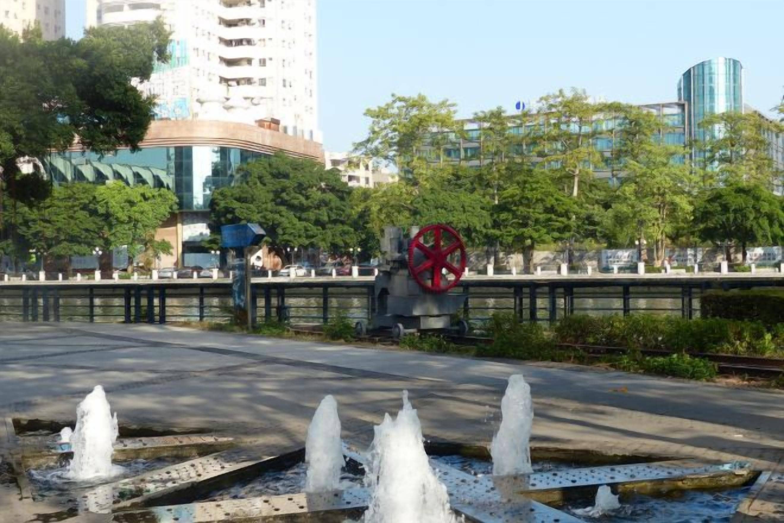 Una fuente con chorros de agua, una rueda decorativa roja y un edificio moderno al fondo.