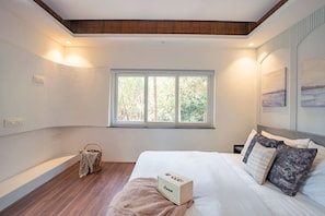 Une chambre avec un grand lit, une fenêtre donnant sur des arbres, une table de chevet et une lampe.