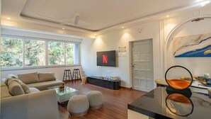 Een moderne woonkamer met een bank, poufs, een glazen salontafel, een aan de muur bevestigde televisie en een schilderij.