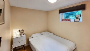 A small, neatly arranged bedroom with a single bed, a bedside table with a lamp, and a window with a view of trees.