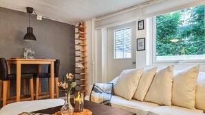 A living room with a wooden table, a sofa, a bookshelf, and a window with a view of greenery.