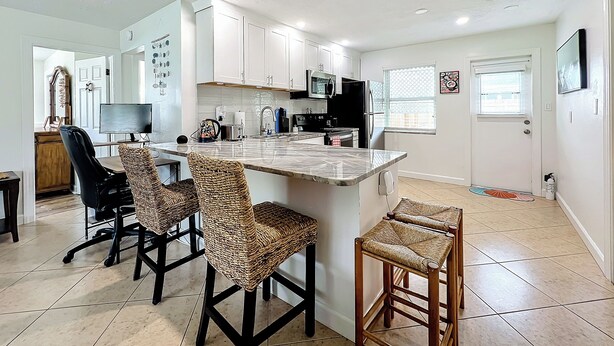 A modern kitchen with white cabinets, a large island with bar stools, and a dining area with a chair and desk.