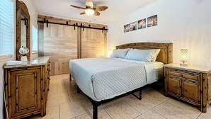 A bedroom with a bed, wooden headboard, bedside tables, a ceiling fan, and a wall-mounted TV.