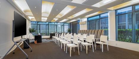 Un salón de conferencias con una pantalla grande, filas de sillas y amplias ventanas que ofrecen vistas al exterior.