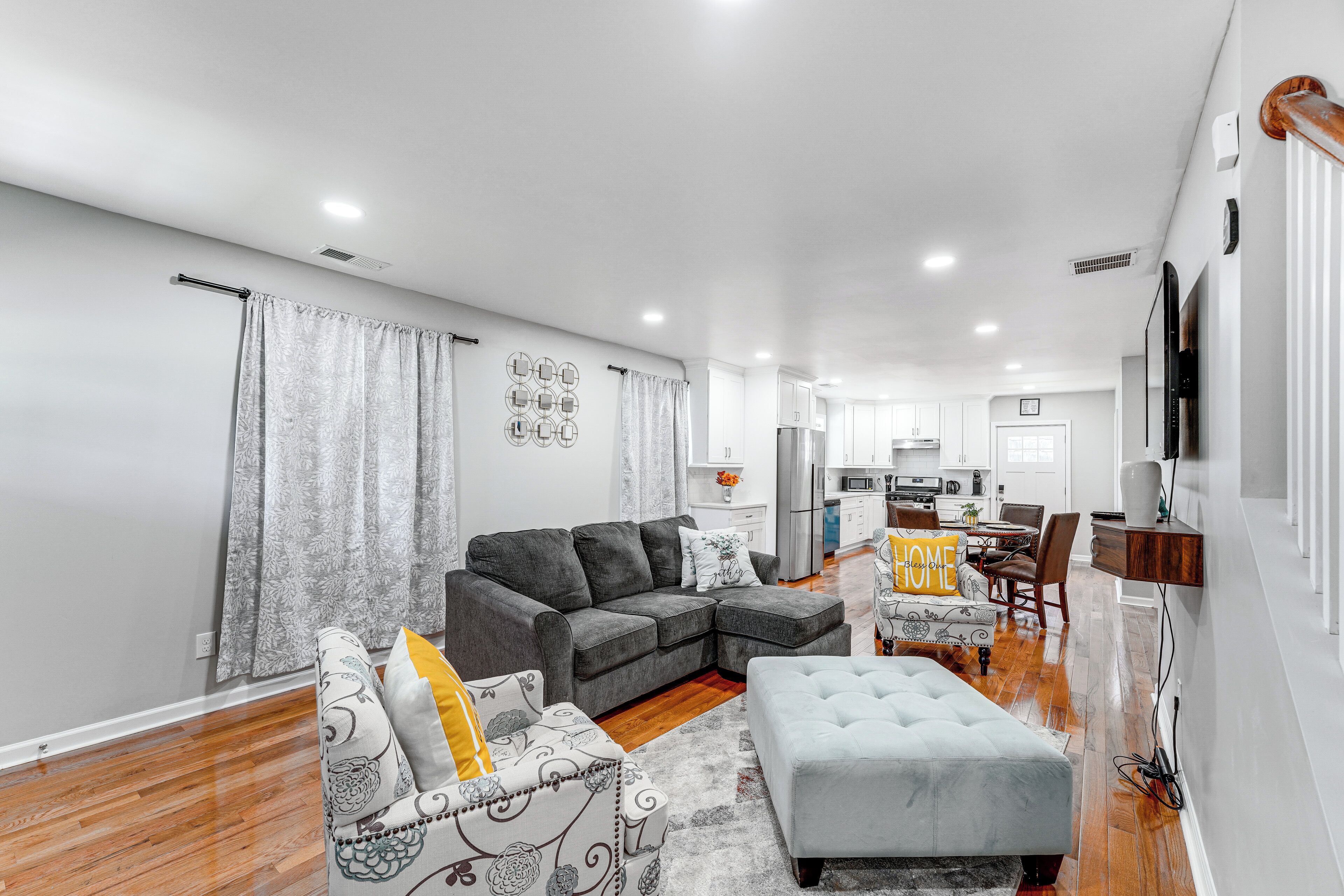 A modern living room with a grey sectional sofa, a patterned armchair, and a grey ottoman. A kitchen is visible in the background with white cabinets and a dining area.