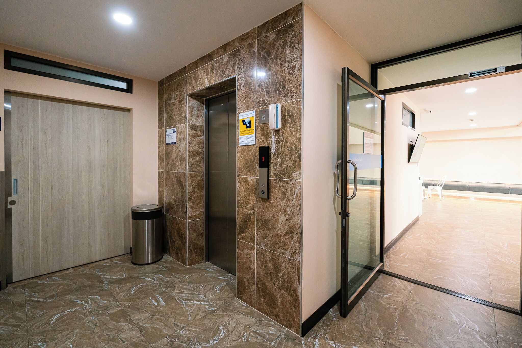 Un ascenseur avec un mur en marbre, une poubelle et une porte en verre donnant sur un couloir.