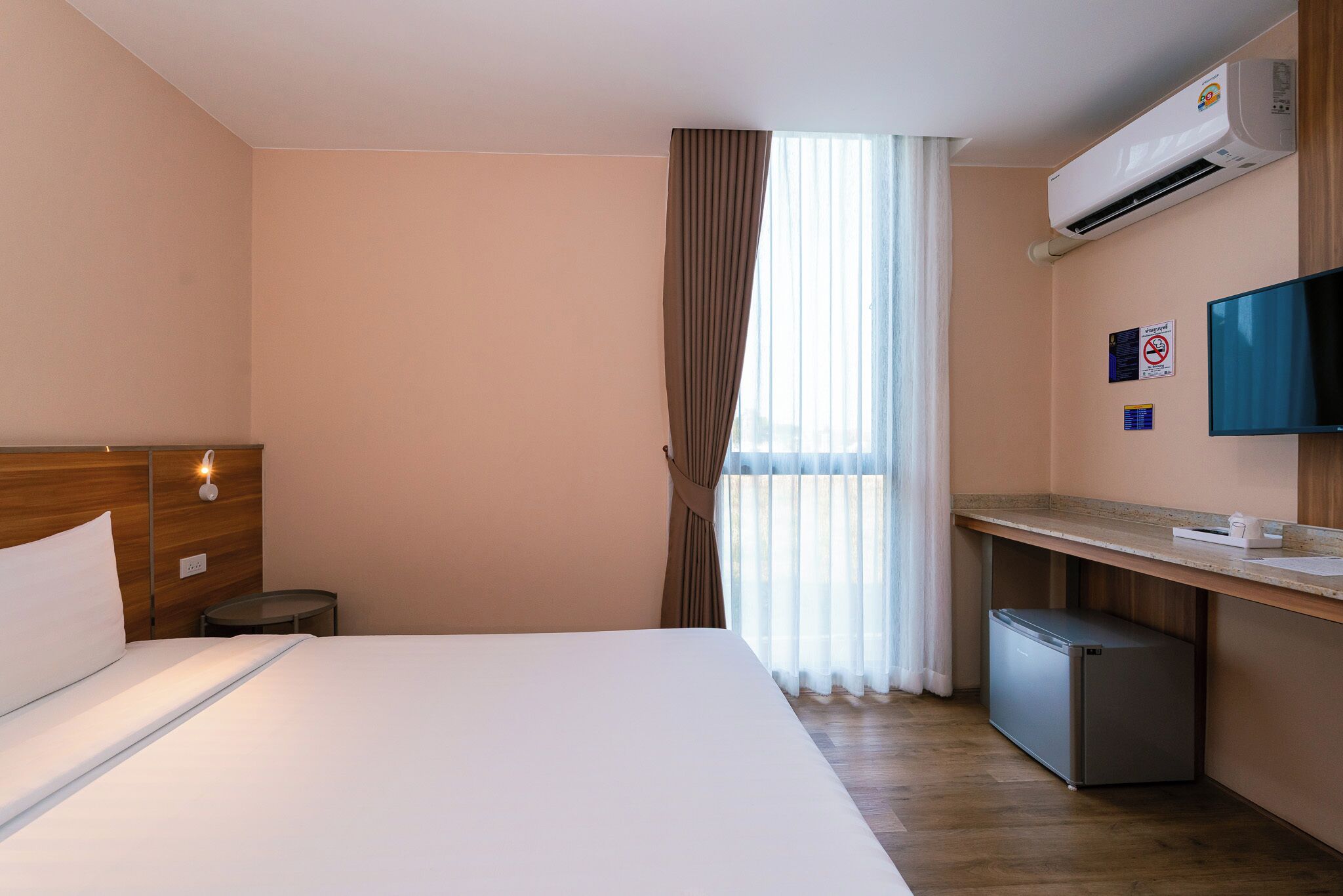 A hotel room with a bed, a desk, a small refrigerator, an air conditioning unit, a television, and a window with curtains.
