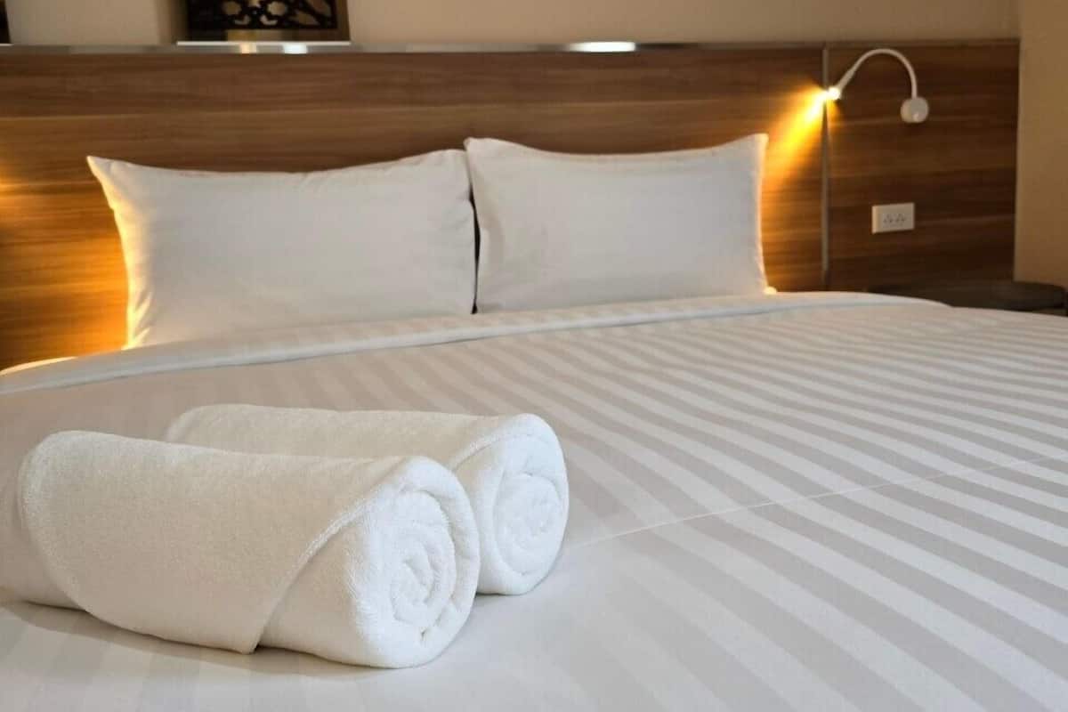 Una cama bien tendida con sábanas blancas y dos toallas enrolladas, cabecera de madera y una lámpara fijada a la pared.