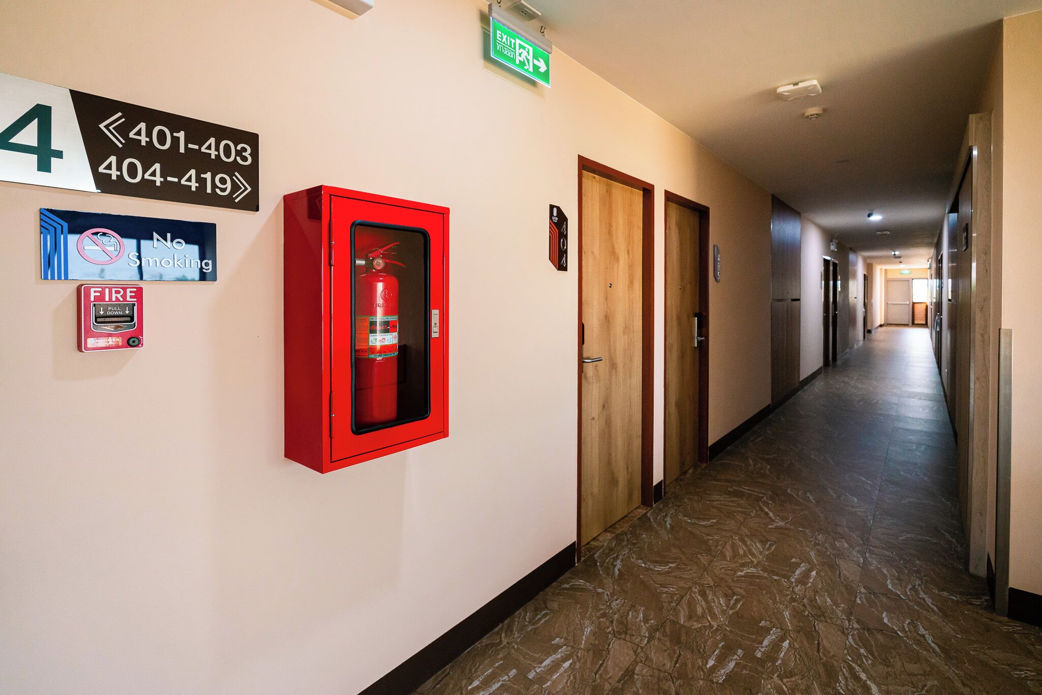 A hotel hallway with a fire extinguisher, emergency exit sign, and a no-smoking sign.