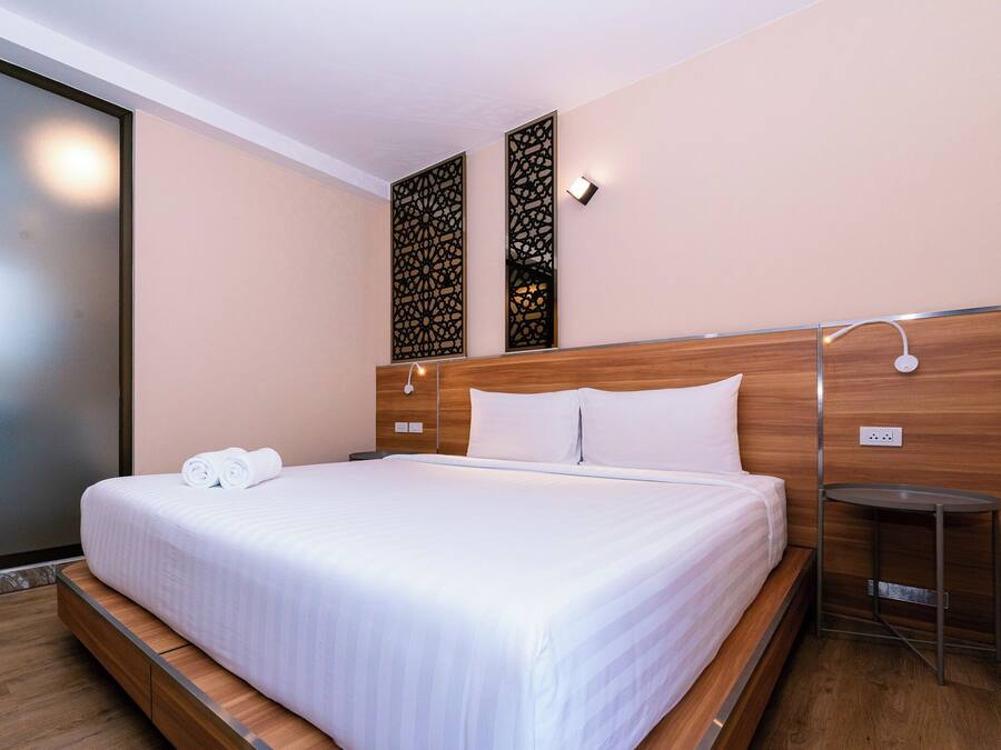 A hotel room with a large bed, a wooden headboard, a nightstand, a wall-mounted lamp, and a decorative panel.