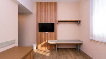 Una habitación pequeña con un armario de madera, un televisor montado en la pared, una banca y una ventana con cortinas.