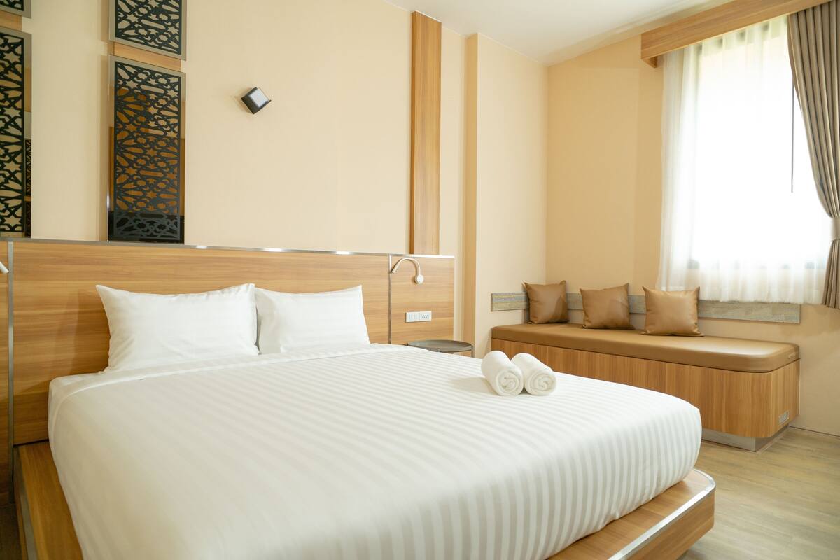 Una habitación de hotel moderna con una cama grande, cabecera de madera, una mesita de noche, un banco empotrado con cojines y una ventana con cortinas.