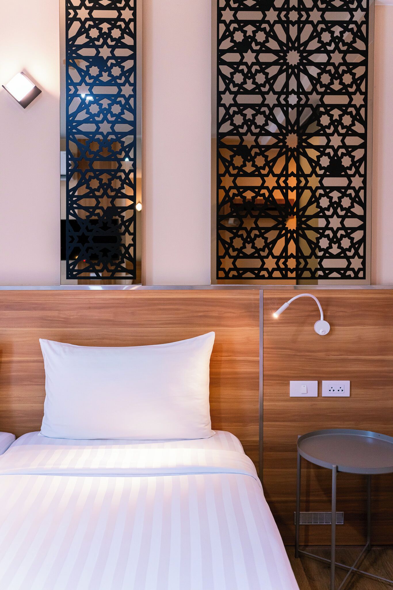 Une chambre d’hôtel comprenant un lit, une table de chevet, une lampe fixée au mur et un paravent décoratif orné de motifs géométriques.