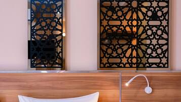 Habitación de hotel con una cama, una mesita de noche, una lámpara de pared y un separador decorativo con motivos geométricos.