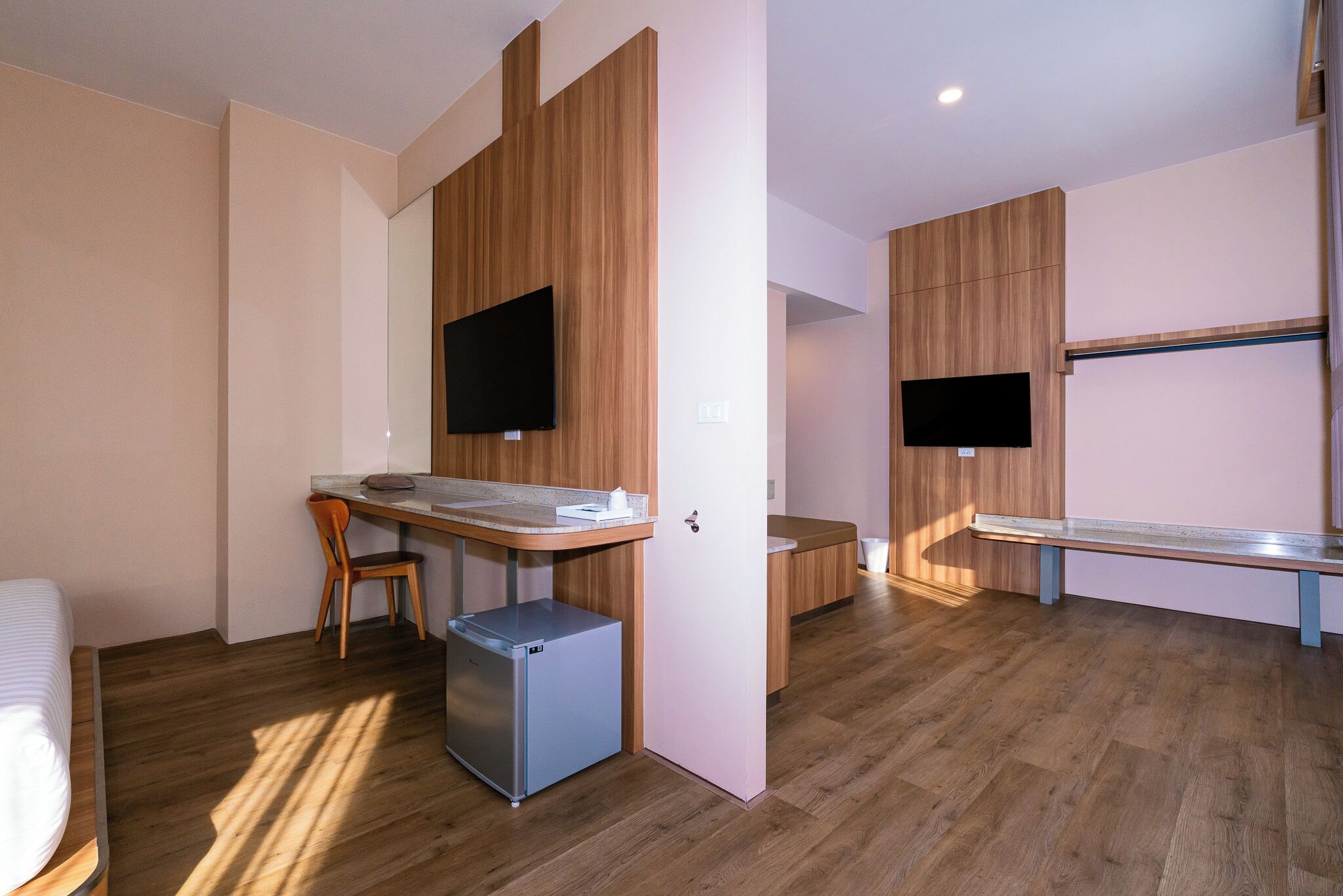 A modern hotel room with a wooden floor, a desk with a chair, a small refrigerator, and two flat-screen TVs mounted on the walls.