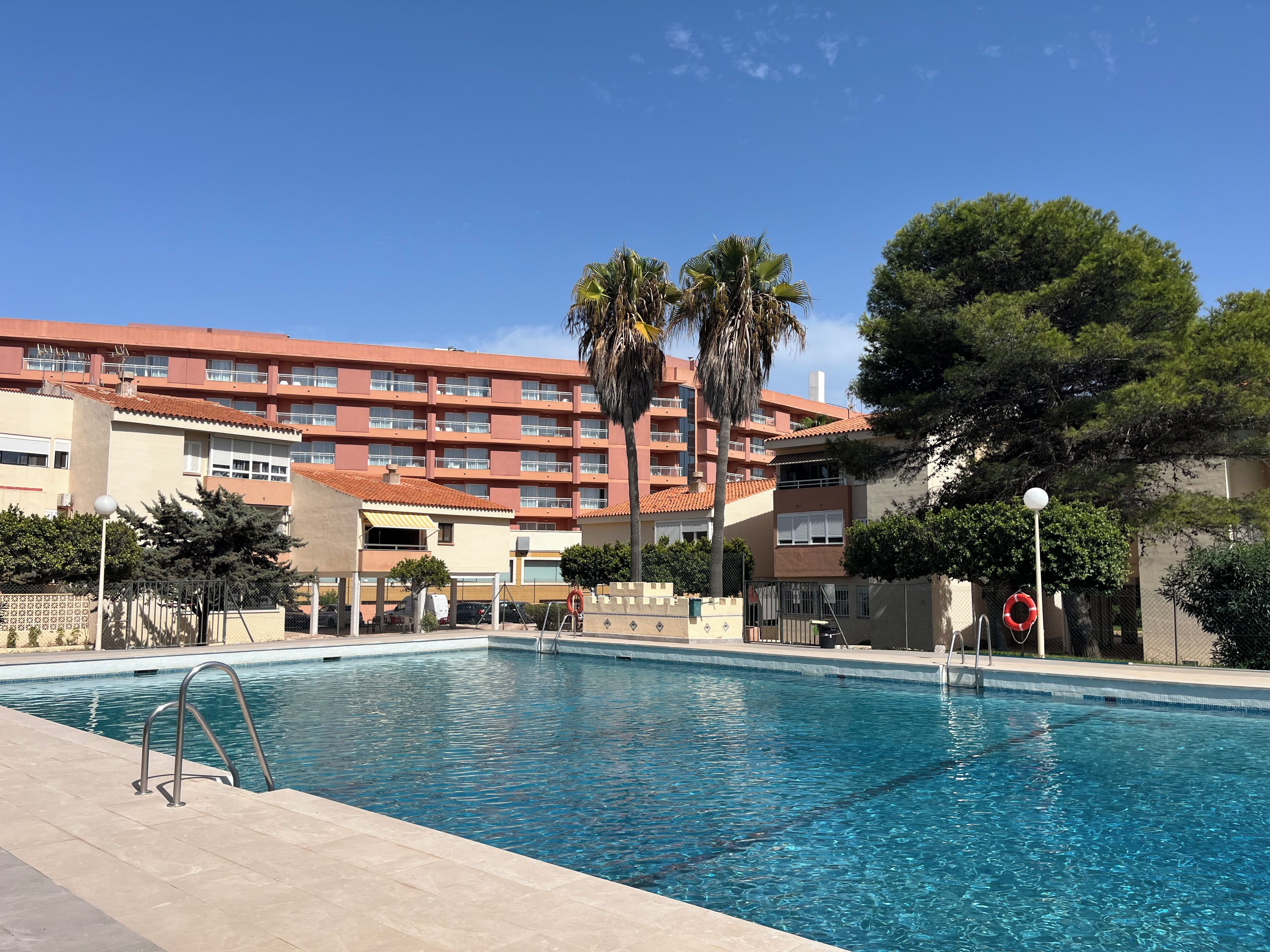 A swimming pool with a clear blue water surface, surrounded by a tiled deck. In the background, there is a multi-story building with balconies and a red-tiled roof.
