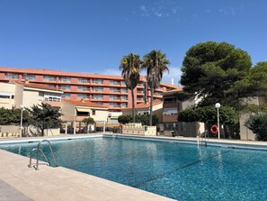 A swimming pool with a clear blue water surface, surrounded by a tiled deck. In the background, there is a multi-story building with balconies and a red-tiled roof.