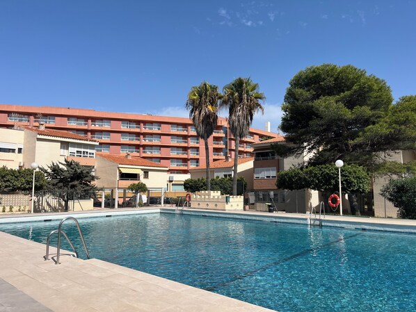 Una piscina con superficie de agua azul cristalina, rodeada por una solana con azulejos. Al fondo, un edificio de varios pisos con balcones y un techo de tejas rojas.