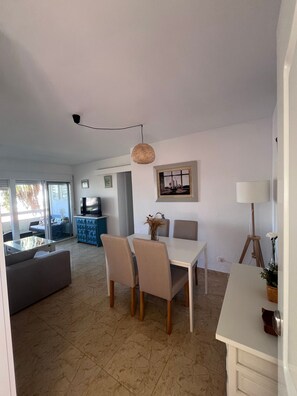A spacious living area with a dining table, chairs, and a sofa. There is a television and a decorative vase on the table.