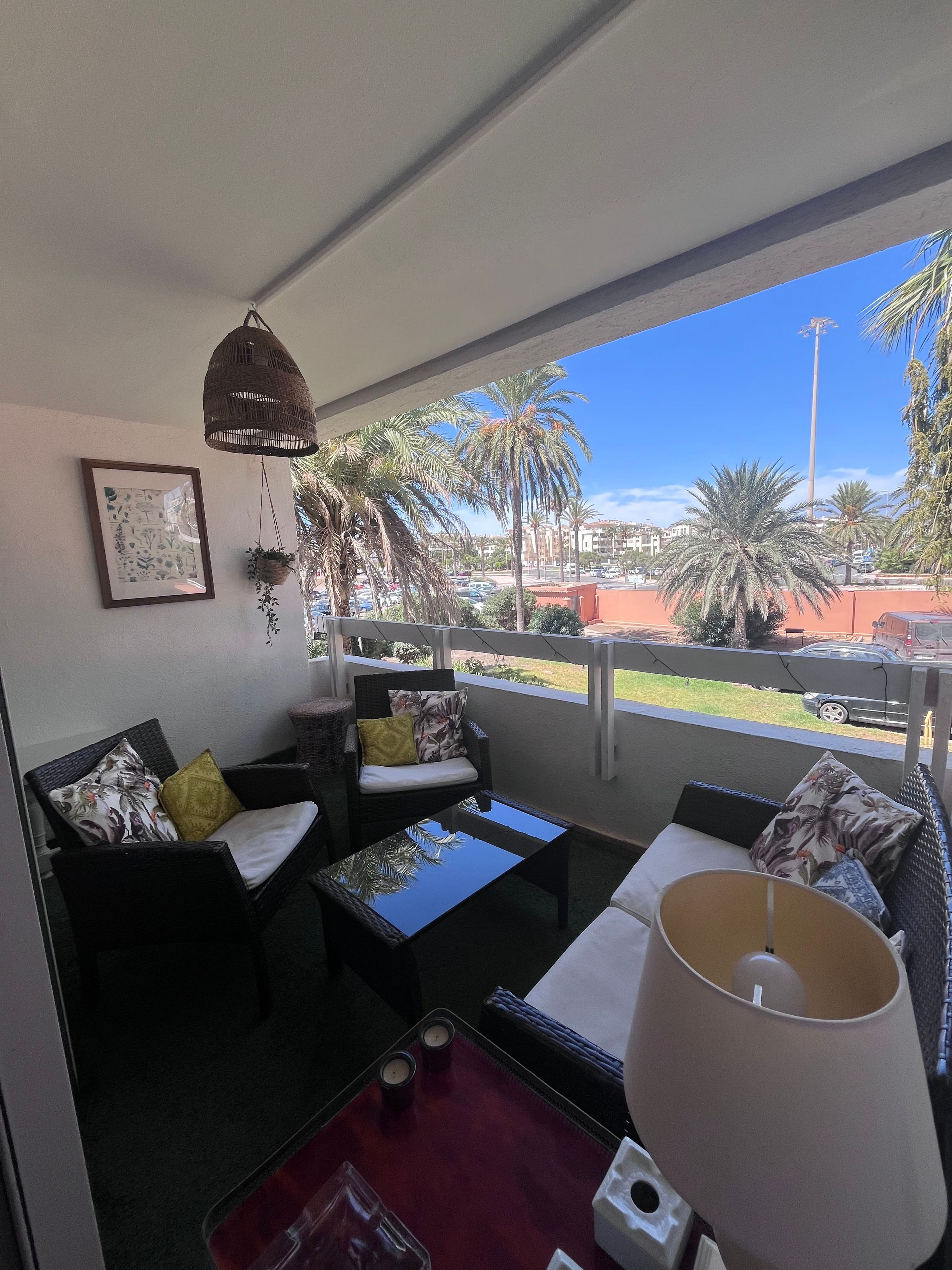 A balcony with wicker furniture, a framed picture, and a view of palm trees and buildings.