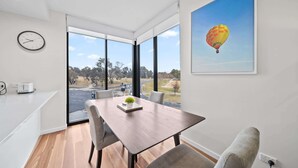 A modern dining area with a wooden table, grey upholstered chairs, a clock on the wall, and a hot air balloon painting. - Inner-City 2-Bed with Large Balcony, Garden & Gym (Campbell)